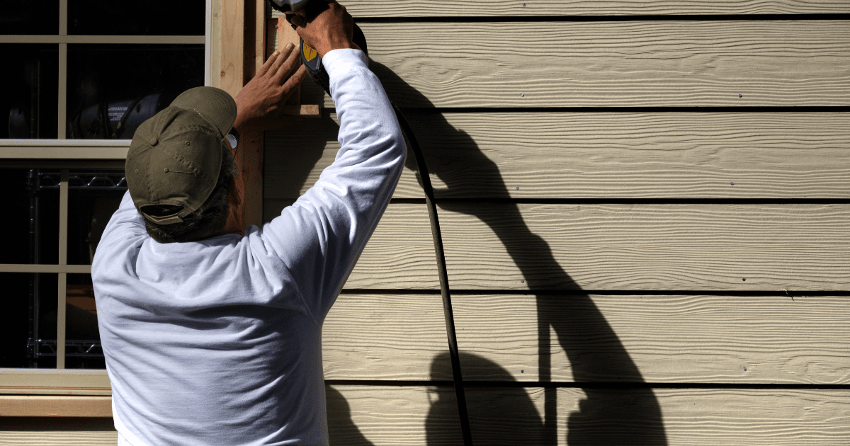 A person in a white shirt uses a power tool on beige siding, casting a distinct shadow. The scene conveys focus and craftsmanship in home improvement.