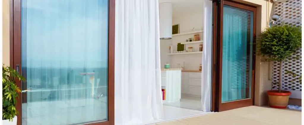 Sliding glass door with sheer curtains open to a bright kitchen interior. Wooden frames, potted plant, and ocean view create a serene, airy atmosphere.