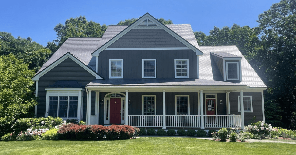 A two-story gray house with a red door, white trim, and a wraparound porch surrounded by manicured greenery under a clear blue sky.