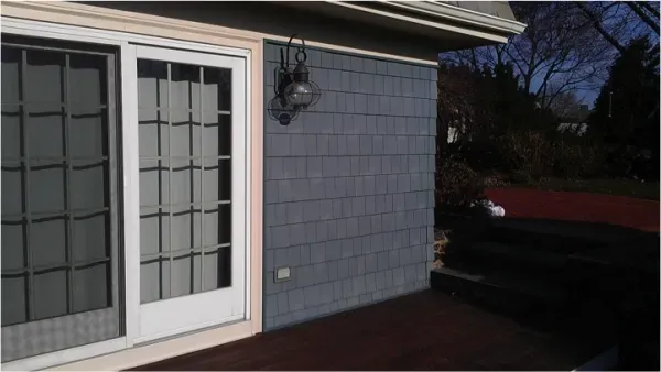 A sunlit, modern gray house exterior features a glass sliding door with grid patterns, a wall lamp, and adjacent dark steps leading to a paved area.
