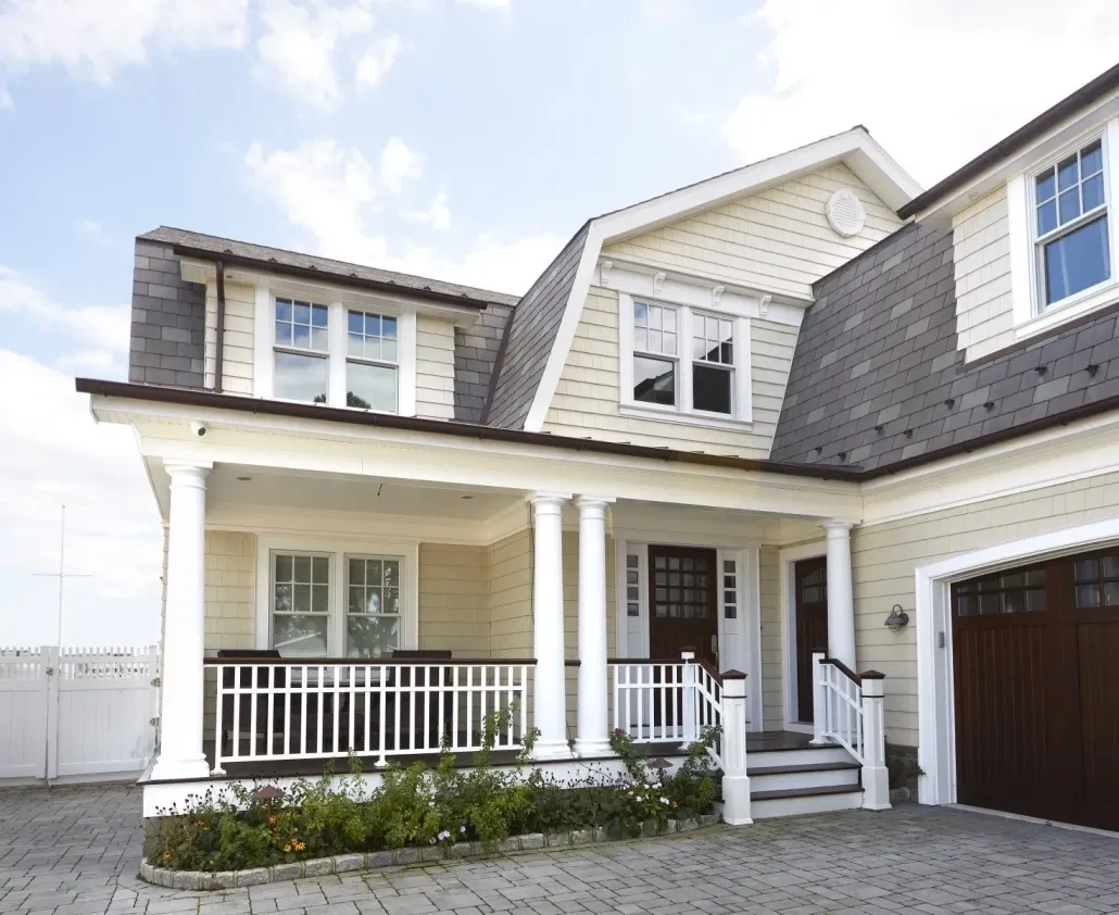 A large beige house with a gable roof, white trim, and dark shingles. It features a spacious porch with white pillars and a neatly paved driveway.