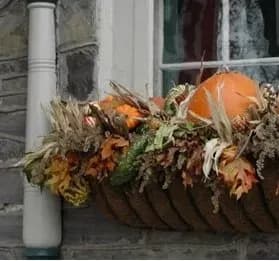 Autumn-themed window box with a pumpkin, dried corn husks, leaves, and gourds against a stone wall backdrop, conveying a rustic, cozy feel.