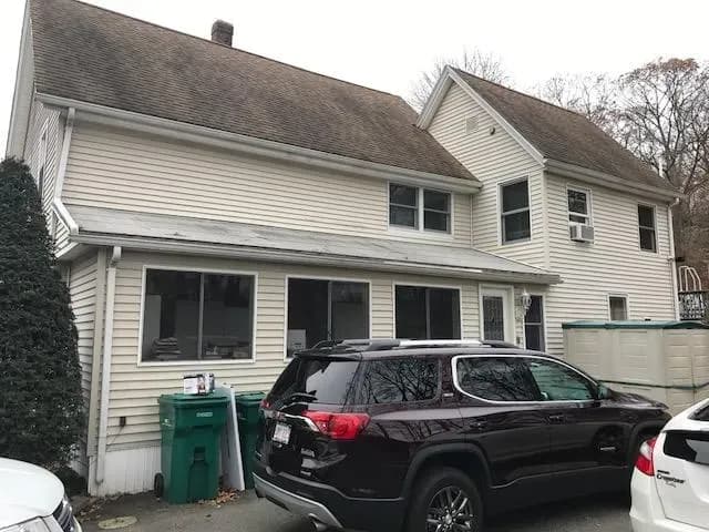 A two-story beige house with a steep, brown roof. A dark SUV is parked in front, alongside green trash bins. Overcast sky and leafless trees.