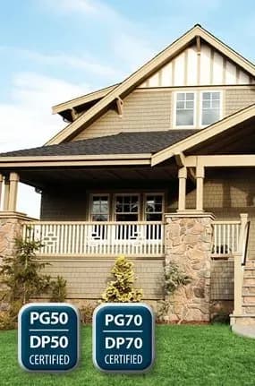 A modern craftsman-style home with stone columns and beige siding under a blue sky. Two badges indicate PG50/DP50 and PG70/DP70 certifications.
