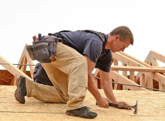 A worker in a navy shirt and khaki pants, with a tool belt, kneels on a roof, focused and hammering nails into the wooden surface.