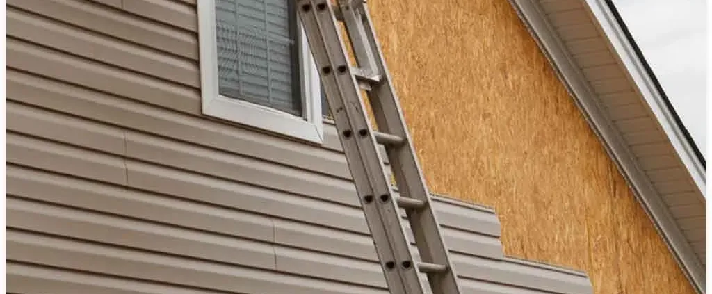 A ladder leans against a house with partially installed siding; part is still wooden. A window with closed blinds is visible. The mood is neutral.