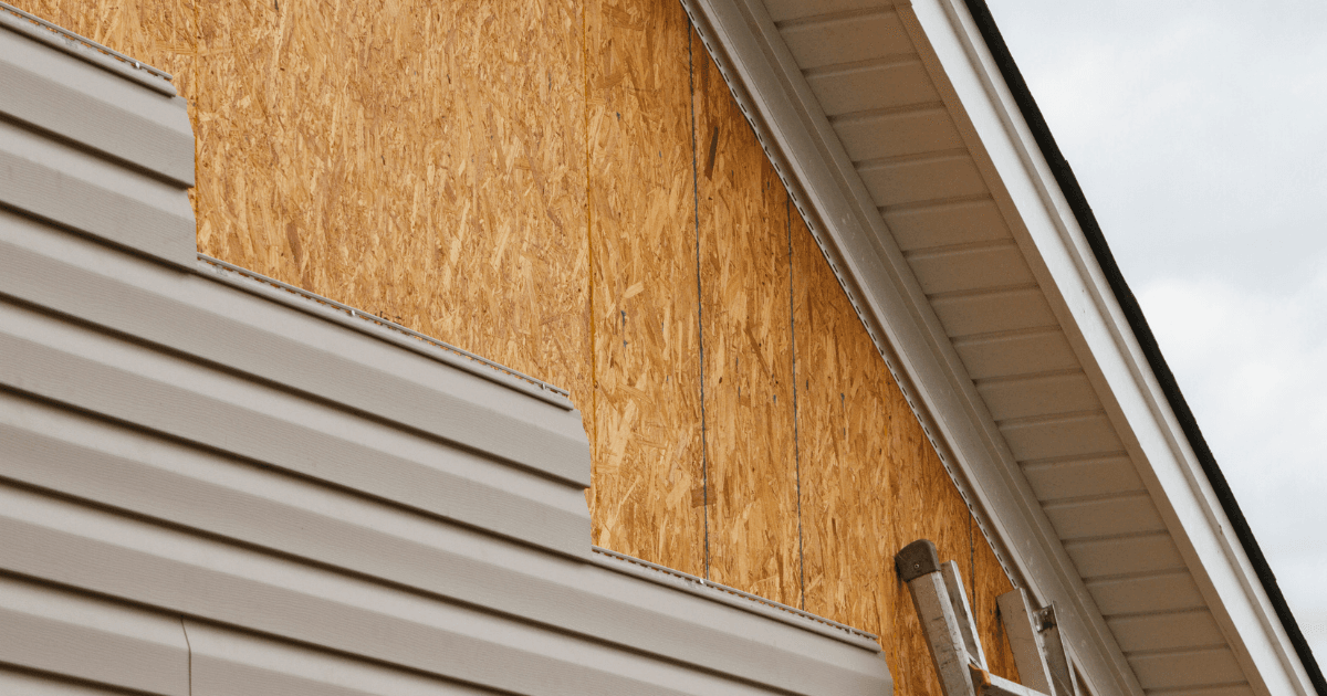 A house under renovation, showing partially installed beige vinyl siding on a wooden exterior. A ladder leans against the building under a cloudy sky.