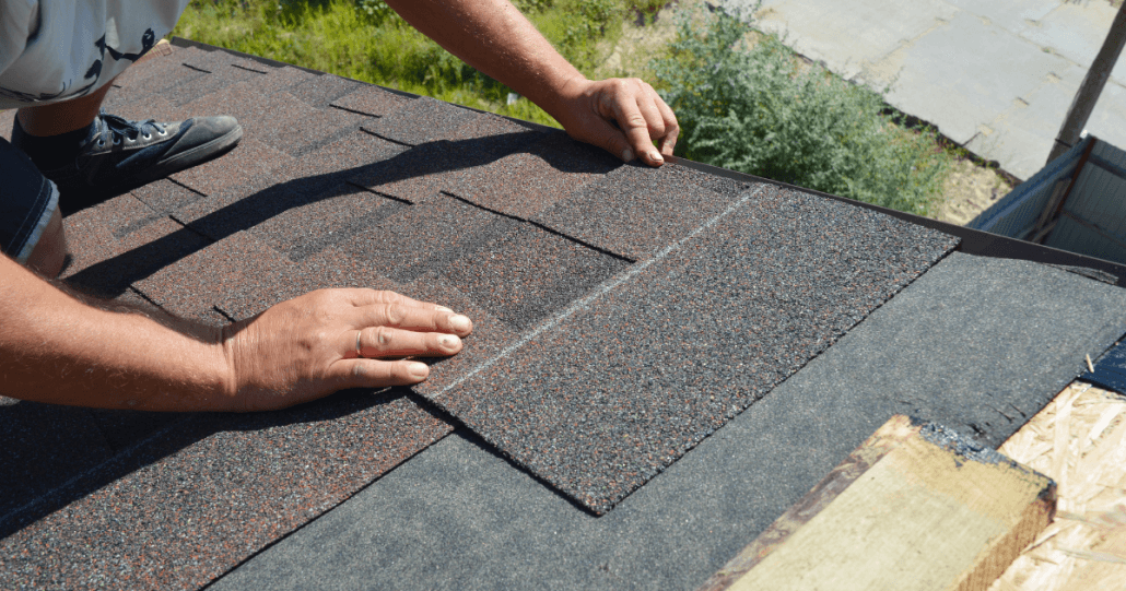 A close-up of a person installing asphalt shingles on a roof. Hands carefully align the dark shingles, conveying precision and craftsmanship.