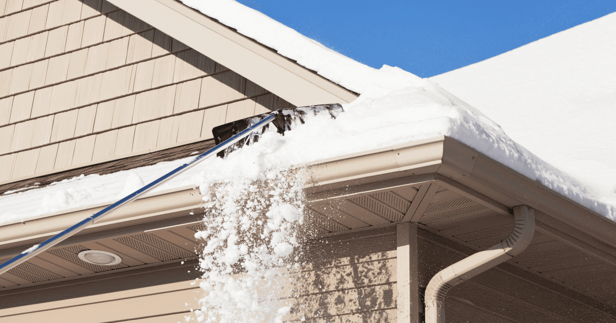 A person uses a roof rake to clear thick snow off a steep rooftop under a bright blue sky. Snow cascades down, highlighting winter maintenance.
