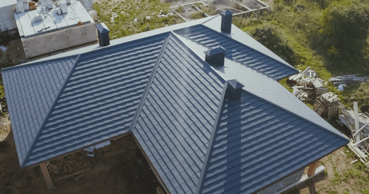 Aerial view of a house featuring a large, blue metal roof with multiple peaks and chimneys. Surrounding landscape includes grass and construction materials.