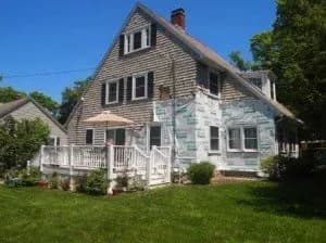 Gabled house with a partly renovated exterior, white siding on one side, shingles on the other. A white porch and green lawn under a clear blue sky.