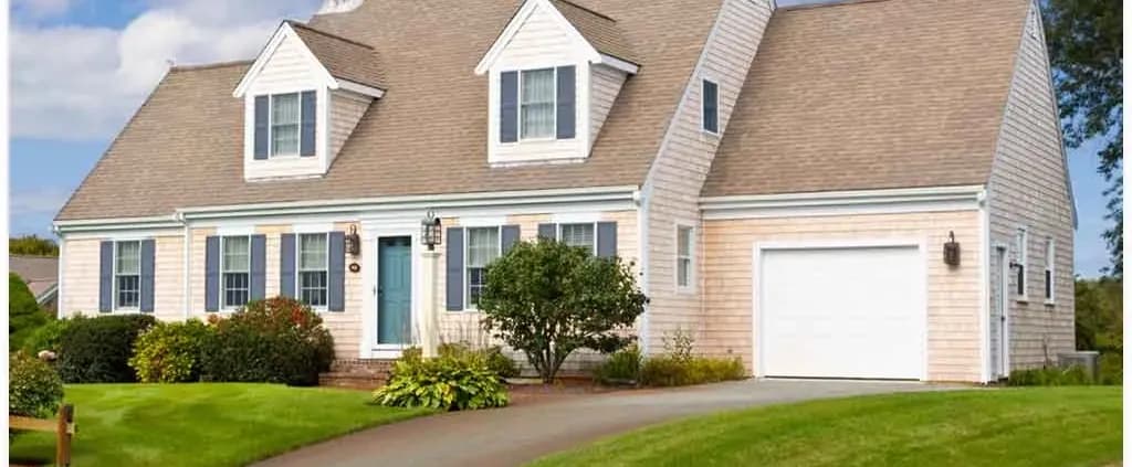 A charming Cape Cod-style house with blue shutters, a teal door, and a dormer window. It sits amid a well-kept lawn with shrubs, under a bright sky.