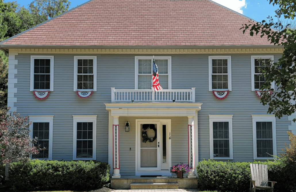 A two-story gray house with a red roof, adorned with American flags and patriotic bunting. A porch displays a wreath and potted flowers, creating a festive vibe.