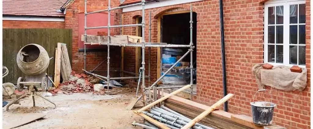 Red brick house under renovation with scaffolding, a cement mixer, and scattered building materials. The scene conveys construction progress.