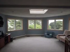 Spacious, softly lit room with five large windows showing lush greenery. Contains a blue armchair, beige sofa, and wooden cabinet, exuding a serene ambiance.