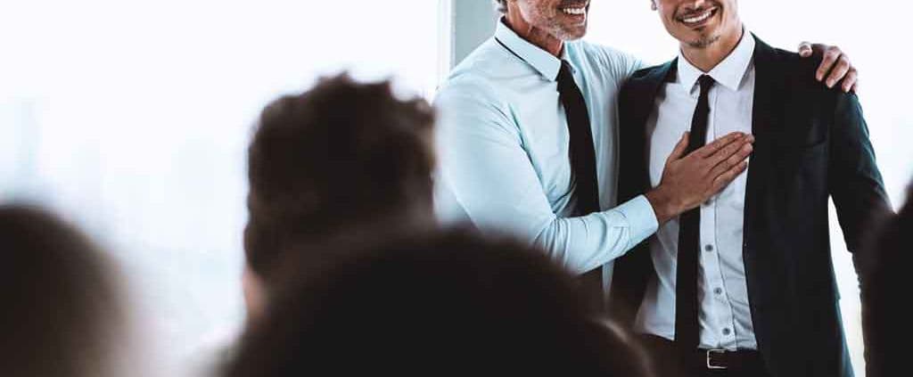 Two men in formal attire stand together, smiling warmly. One has his arm around the other, conveying a sense of camaraderie and celebration.