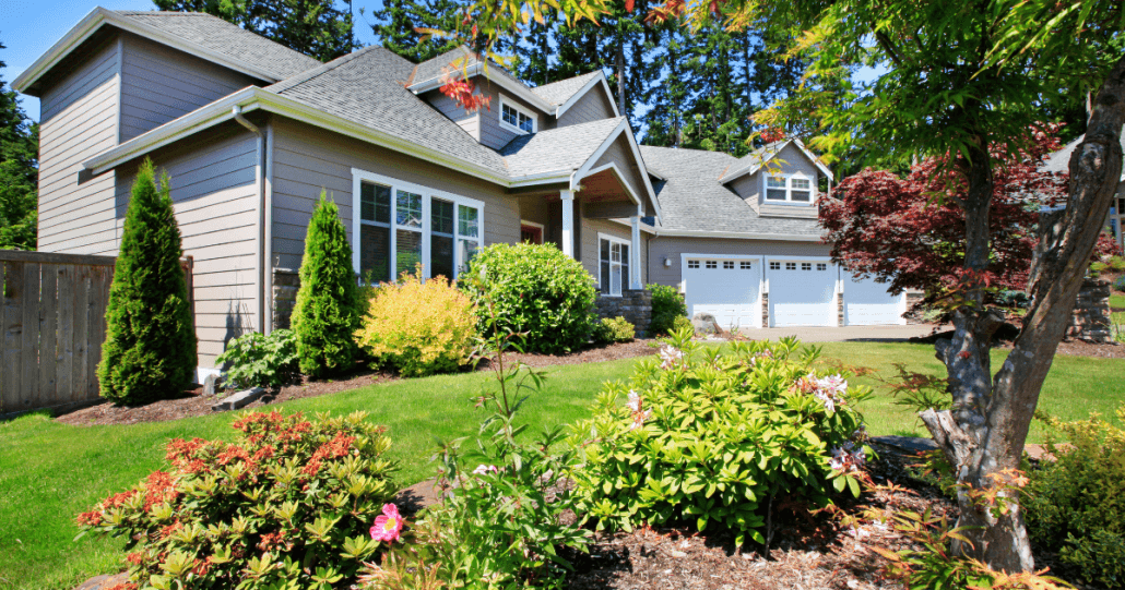 A charming suburban house with a gray exterior and a two-car garage sits amid a lush, well-maintained garden. Bright flowers and shrubs line the path.