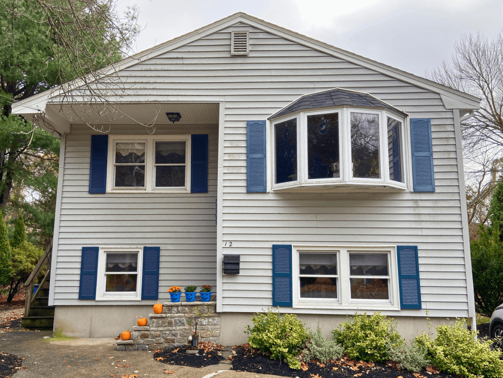 Gray two-story house with blue shutters, a bay window, and pumpkins on stone steps. Surrounded by trees and shrubs, the scene feels quaint and autumnal.