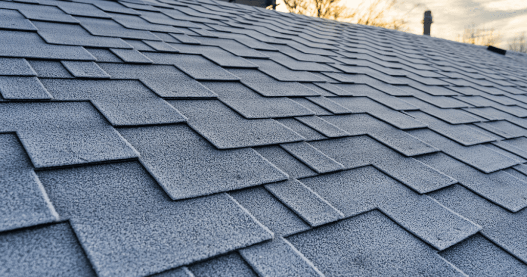 Close-up of a frost-covered shingle roof at sunrise. The overlapping shingles create a textured pattern, evoking a cold, early morning atmosphere.