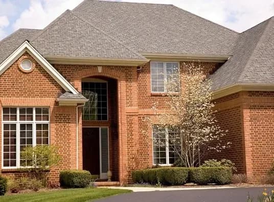 A two-story brick house with a gabled roof, large windows, a tall arched entryway, surrounded by manicured bushes and a flowering tree.