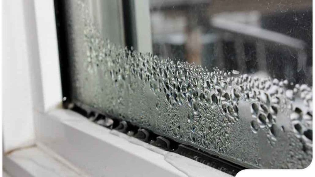 Condensation droplets on a window pane, with a blurry view of an urban landscape outside. The scene feels cool and humid, suggesting a rainy day.