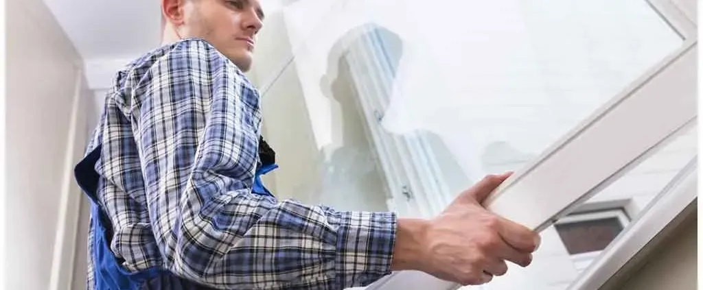 A man in a plaid shirt is carefully installing a window inside a bright room. His expression is focused, reflecting concentration and precision.