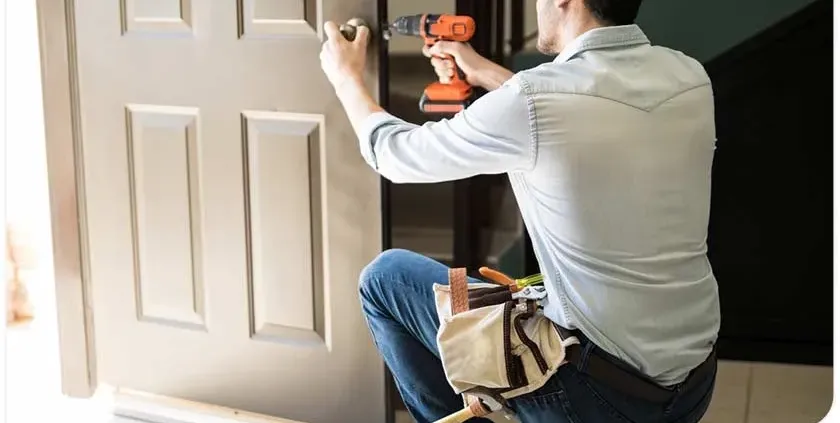 A person in a light shirt and jeans kneels, using an orange drill on a beige door. They have a tool belt, suggesting home repair or installation work.