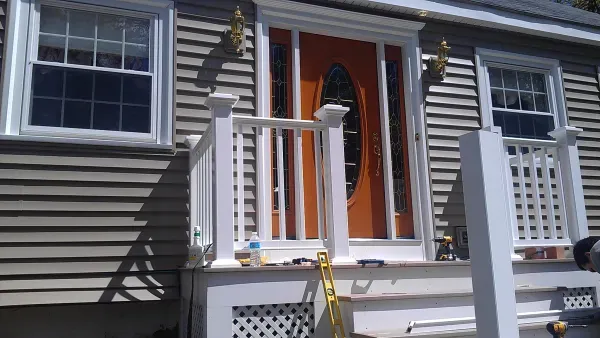 A house entrance featuring a bold orange door with a decorative oval glass pane, flanked by two windows. A small porch and white railings are in the foreground, with a yellow ladder leaning against them, indicating ongoing work. The scene is well-lit with sunlight, creating a bright, welcoming atmosphere.