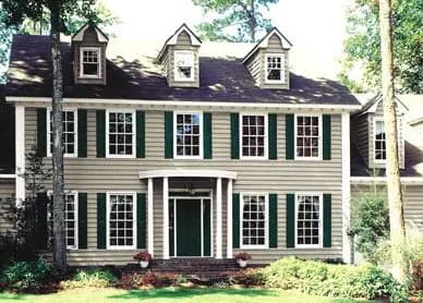 Two-story house with green shutters and beige siding. Symmetrical facade features multiple windows, dormers, and a central green door. Surrounded by trees.