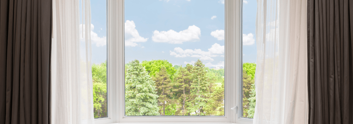 A large window with sheer curtains frames a scenic view of lush green trees and a bright blue sky with fluffy clouds, creating a serene atmosphere.