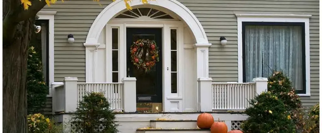 Front porch of a home decorated for autumn. Two pumpkins sit by the steps, and a wreath of fall leaves hangs on the door, evoking a cozy, festive atmosphere.