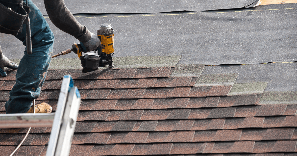 Worker using a nail gun to install asphalt shingles on a roof. The scene shows a close-up of the process, focusing on precision and craftsmanship.