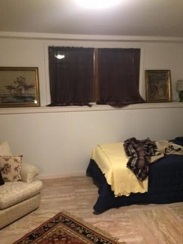 A cozy bedroom with light walls featuring a bed with yellow and dark bedding, two framed paintings, a plush chair, and a carpet, conveying warmth and comfort.