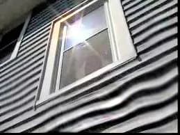 Sunlight reflects off a window on a house with warped, wavy siding, creating a bright spot. The scene suggests intense heat or distortion effects.