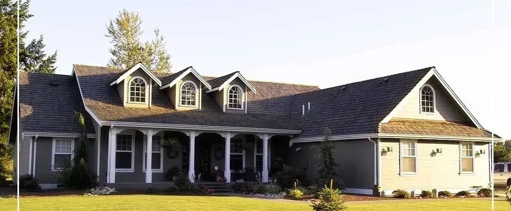A charming two-story gray house with white trim, featuring three gabled dormer windows and a porch, set in a well-kept yard with lush green grass.