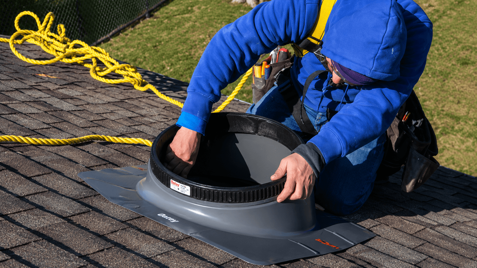 A person in a blue hoodie and safety harness installs a solar tube on a rooftop. They are focused on their work, with tools at their waist and a yellow safety rope secured behind them.
