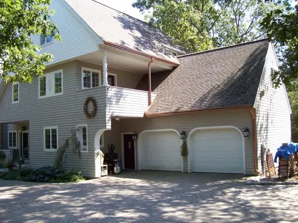 Two-story suburban house with tan siding, a double garage, and a welcoming front porch. The scene is serene, with trees and sunlight enhancing its coziness.