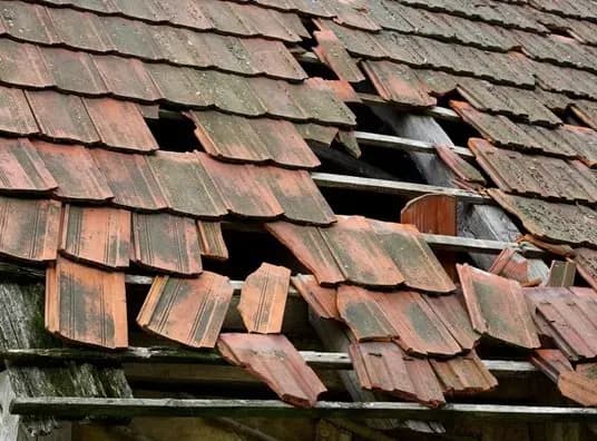 Damaged roof with missing and broken red tiles exposing wooden beams underneath. The aged, weathered materials evoke neglect and decay.