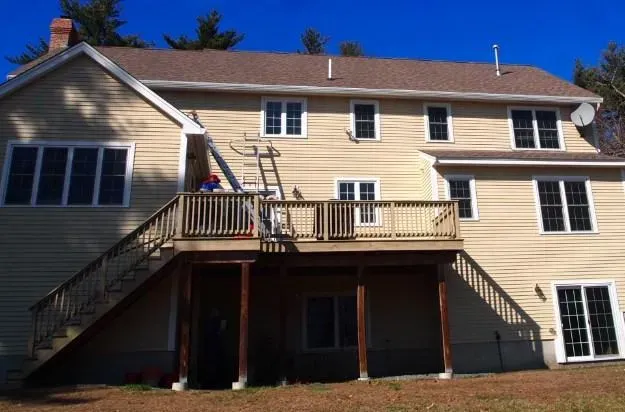 A beige two-story house with a sloped roof, large deck, and outdoor stairs. Clear blue sky above; a ladder rests against the deck railing.