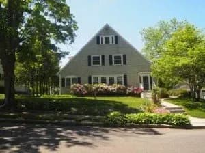 A-frame gray house with black shutters, flanked by lush greenery and blooming bushes. Sunlit yard, blue sky, and trees create a serene suburban setting.