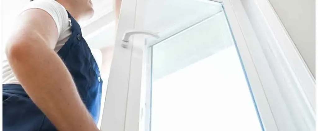 A person in work overalls installs a white-framed window, focused and intent. Bright natural light streams through, creating a calm atmosphere.