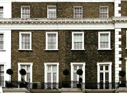 Historic brown brick building with white-framed windows and black wrought-iron balconies, adorned with round topiary trees, conveys elegance.