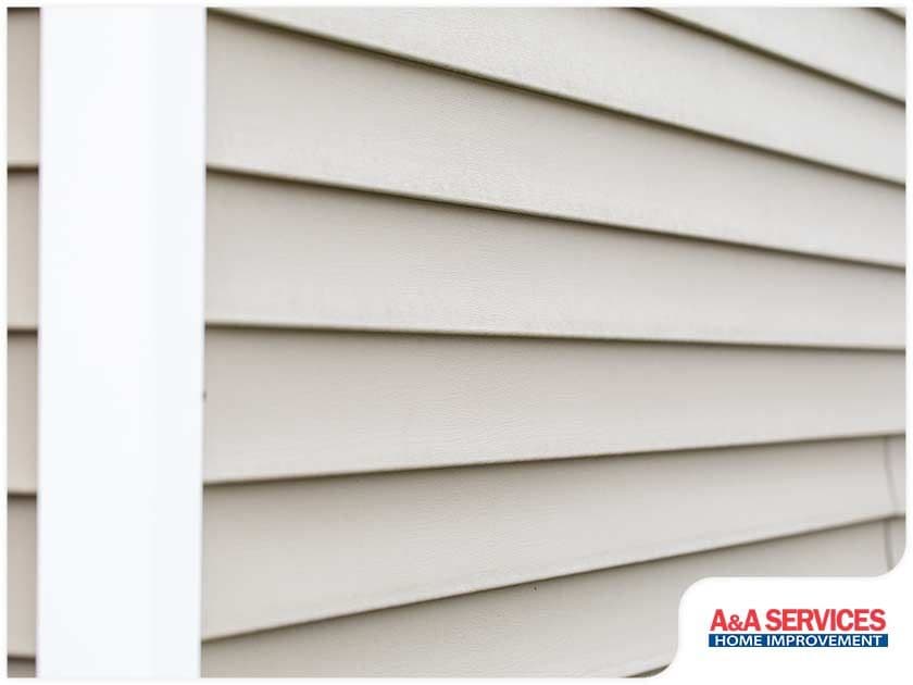Close-up of beige vinyl siding on a house, angled to show texture and alignment. A vertical white trim is visible. A&A Services Home Improvement logo at the corner.