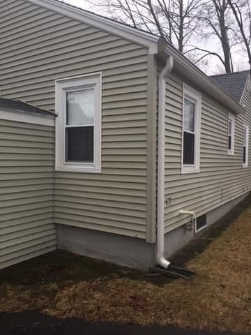 Single-story house corner with beige vinyl siding, two white-framed windows, and a downspout. Sparse winter trees in the background. Overcast sky.