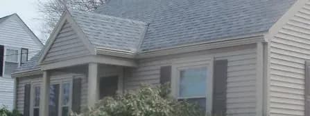 A cozy, beige, single-story house with a gabled roof and large front windows, partially obscured by lush greenery, evoking a welcoming ambiance.