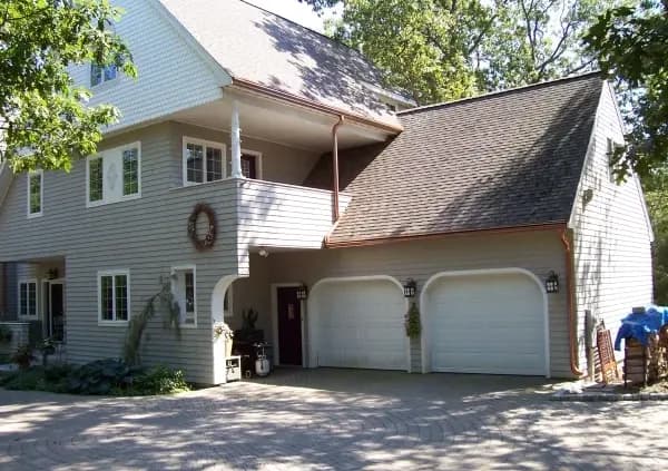 Two-story suburban home with gray siding, a two-car garage, and a driveway. A wreath adorns a column. Trees surround the house, adding a serene feel.
