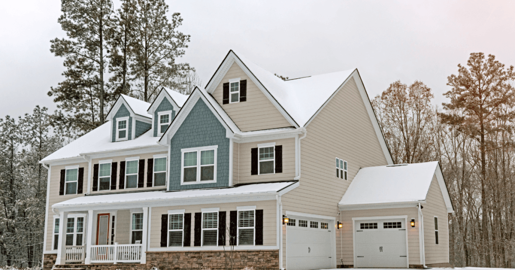 A large, two-story suburban house with blue and beige siding sits surrounded by snow and tall trees, conveying a serene winter atmosphere.