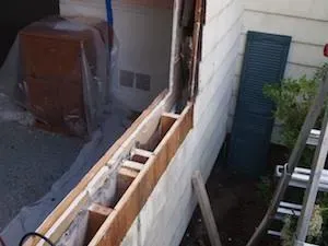 A partially dismantled wall with exposed wooden beams and insulation, alongside a covered piece of furniture. A ladder leans against the house, suggesting renovation.