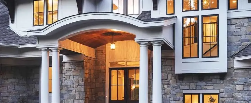 Elegant stone house at dusk with warm lit windows, large arched entrance, two-story columns, and a mix of traditional and modern architectural features.