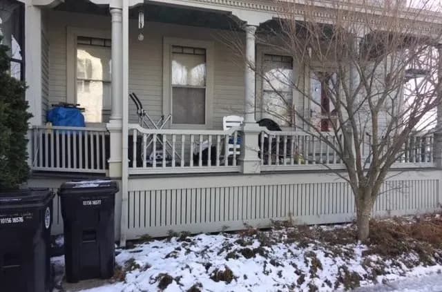 A snowy porch scene with two black garbage bins in the foreground. The porch has wooden railings and scattered outdoor items, creating a cluttered appearance.